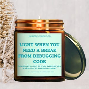 May include: A candle in a brown jar with a gold lid, labeled "LIGHT WHEN YOU NEED A BREAK FROM DEBUGGING CODE." The candle is next to a gold star and small Christmas trees. The text on the label reads "Infused with a hint of stack overflow and a whole lot of existential errors."