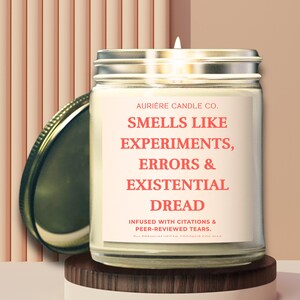 May include: A glass jar candle with a silver lid, labeled "AURIERE CANDLE CO." and the text "SMELLS LIKE EXPERIMENTS, ERRORS & EXISTENTIAL DREAD." The candle sits on a wooden and white platform. The background is a peach color.