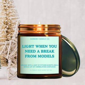 May include: A brown glass candle jar with a gold lid, featuring the text "LIGHT WHEN YOU NEED A BREAK FROM MODELS." The candle is surrounded by small Christmas trees and a star-shaped ornament. The candle is made with vegan coconut soy wax.