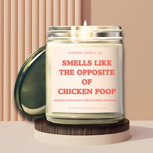 May include: A glass jar candle with a silver lid, placed on a wooden stand. The candle has a label that reads "Smells Like The Opposite Of Chicken Poop." The background features a peach-colored, vertical striped design.