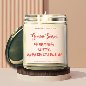 May include: A glass jar candle with a silver lid, labeled "Gemini Season" with the words "Charming, Witty, Unpredictable AF." The candle sits on a wooden and white platform. The background is a light peach color.