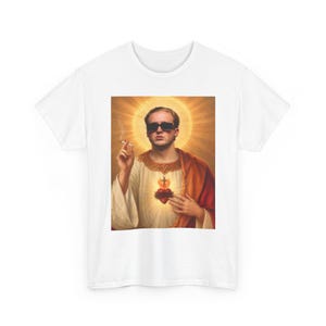 Könnte beinhalten: Ein weißes T-Shirt mit einem Grafikdruck einer Person mit schwarzer Sonnenbrille, bekleidet mit einem cremefarbenen Gewand und einem orangefarbenen Umhang, die eine brennende Zigarette vor einem goldenen Heiligenschein und einem Herzsymbol hält.