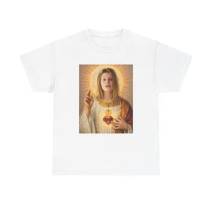 Può includere: Una t-shirt bianca a maniche corte con una stampa grafica centrale che raffigura una persona con un'aureola dorata, che indossa una veste bianca con colletto dorato e tiene una mano sollevata. Un cuore rosso luminoso con croce è visibile sul petto.