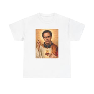 Könnte beinhalten: Ein weißes T-Shirt mit einem Grafikdruck einer Person mit lockigem Haar, die schwarze Kopfhörer um den Hals trägt, in ein weißes Gewand mit roter Schärpe gekleidet ist, eine Zigarette hält und mit einem leuchtenden Heiligenschein dargestellt ist.
