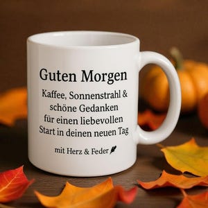 May include: White ceramic mug with the text "Guten Morgen" and other German text. The mug is surrounded by orange and yellow fall leaves and small pumpkins. The mug has a curved handle.