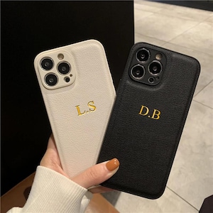 Op de afbeelding: Twee gepersonaliseerde telefoonhoesjes, één witte en één zwarte. De witte hoes heeft de gouden initialen "L.S" en de zwarte hoes heeft de gouden initialen "D.B". Beide hoesjes hebben een textuur die op leer lijkt en een cameramodule.