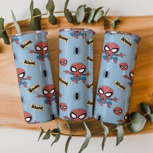 May include: Three light blue insulated tumblers featuring a Spider-Man design. The design includes Spider-Man illustrations, spider icons, and the word "BOOM!" in a comic book style. The tumblers are cylindrical and have a silver rim.
