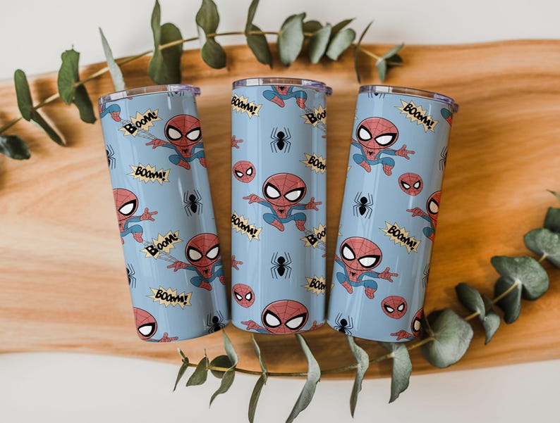 May include: Three light blue insulated tumblers featuring a Spider-Man design. The design includes Spider-Man illustrations, spider icons, and the word "BOOM!" in a comic book style. The tumblers are cylindrical and have a silver rim.