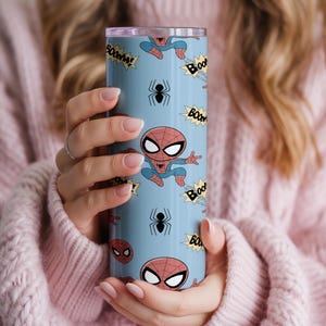 May include: A light blue tumbler featuring a repeating pattern of Spider-Man illustrations, black spiders, and "BOOM!" text bubbles. The tumbler has a silver lid and is held by a person wearing a pink sweater.