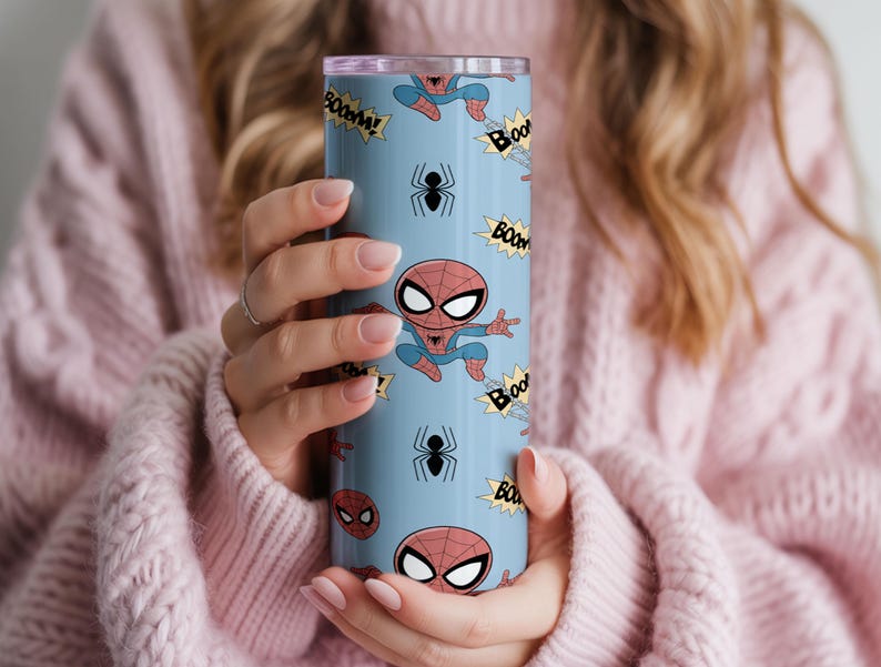 May include: A light blue tumbler featuring a repeating pattern of Spider-Man illustrations, black spiders, and "BOOM!" text bubbles. The tumbler has a silver lid and is held by a person wearing a pink sweater.