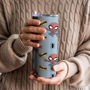 May include: A light blue tumbler featuring a cartoon Spider-Man design. The tumbler has a repeating pattern of Spider-Man figures, "BOOM" text, and black spider illustrations. The tumbler is held by a person wearing a tan sweater.