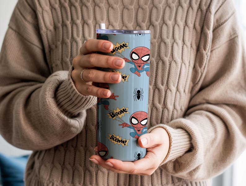 May include: A light blue tumbler featuring a cartoon Spider-Man design. The tumbler has a repeating pattern of Spider-Man figures, "BOOM" text, and black spider illustrations. The tumbler is held by a person wearing a tan sweater.