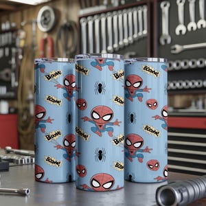 May include: Three cylindrical tumblers with a light blue background featuring a repeating pattern of the Spider-Man character, spider icons, and the word "Boom!" in a comic book style. The tumblers are displayed on a metal surface.