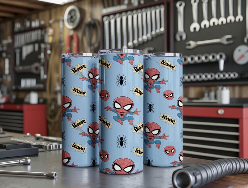 May include: Three cylindrical tumblers with a light blue background featuring a repeating pattern of the Spider-Man character, spider icons, and the word "Boom!" in a comic book style. The tumblers are displayed on a metal surface.