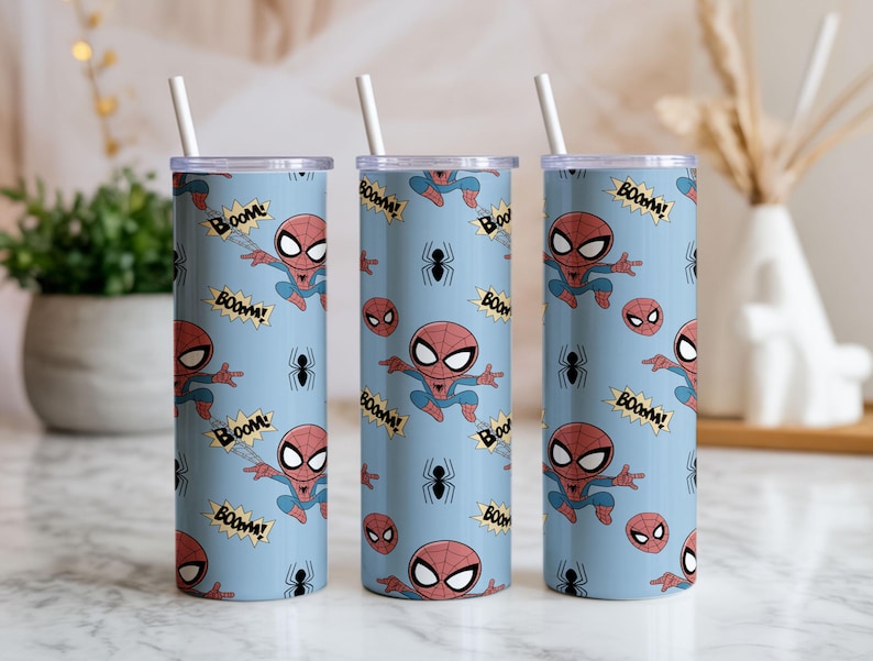May include: Three cylindrical tumblers with a light blue background, featuring a repeating pattern of Spider-Man illustrations, spider icons, and "BOOM!" speech bubbles. Each tumbler has a white straw.