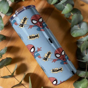 May include: A light blue tumbler featuring a repeating pattern of the Spider-Man character in action poses, along with the word "BOOM!" in yellow starbursts and black spider icons. The tumbler has a silver lid.