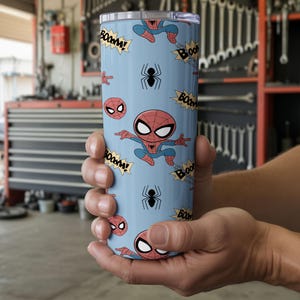 May include: A light blue tumbler featuring a repeating pattern of Spider-Man cartoon illustrations, spider icons, and "BOOM!" text bubbles. The tumbler is cylindrical and appears to be made of stainless steel.