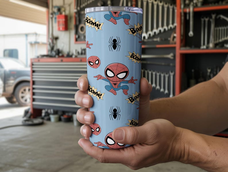 May include: A light blue tumbler featuring a repeating pattern of Spider-Man cartoon illustrations, spider icons, and "BOOM!" text bubbles. The tumbler is cylindrical and appears to be made of stainless steel.