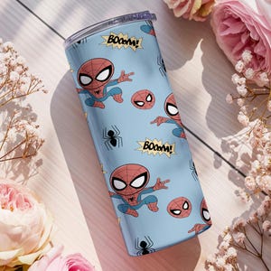 May include: A light blue tumbler featuring a repeating pattern of the Spider-Man character, spider emblems, and the word "BOOM!". The tumbler has a clear lid and is surrounded by pink roses and white flowers.