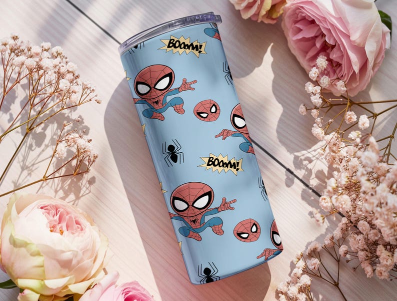 May include: A light blue tumbler featuring a repeating pattern of the Spider-Man character, spider emblems, and the word "BOOM!". The tumbler has a clear lid and is surrounded by pink roses and white flowers.