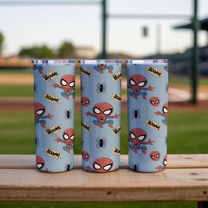 May include: Three stainless steel tumblers with a light blue background. The tumblers are decorated with a repeating pattern of the Spider-Man character, spider webs, and the word "Boom!". The tumblers are sitting on a wooden surface.