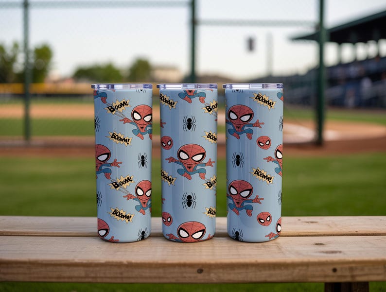 May include: Three stainless steel tumblers with a light blue background. The tumblers are decorated with a repeating pattern of the Spider-Man character, spider webs, and the word "Boom!". The tumblers are sitting on a wooden surface.