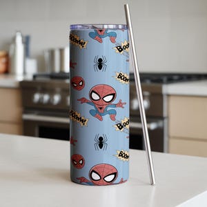 May include: A light blue tumbler with a Spider-Man cartoon pattern. The design includes Spider-Man figures, spider webs, and "BOOM!" text. A silver-colored metal straw is placed next to the tumbler. The tumbler is cylindrical and appears to be made of stainless steel.