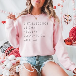 May include: A light pink sweatshirt with long sleeves and the text "1N73LL1G3NC3 15 7H3 4B1L17Y 70 AD4P7 70 CH4NG3" printed in black. The sweatshirt is paired with denim shorts.