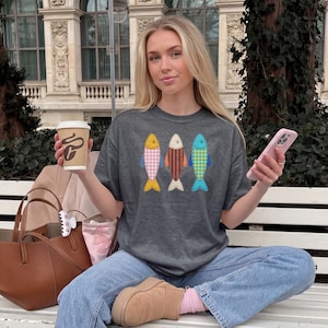May include: A dark gray short-sleeve t-shirt featuring a graphic of three stylized fish in a row. The fish are patterned with pink and white gingham, orange and black vertical stripes, and blue and yellow checkered designs respectively.