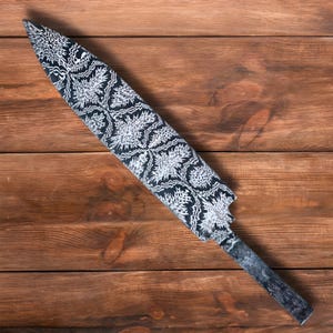 May include: A decorative, long, black and white blade with a detailed floral pattern. The blade has a textured, dark gray handle. The object is set against a wooden background.
