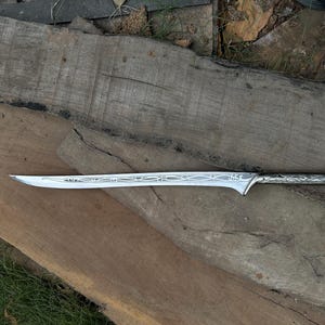 Könnte beinhalten: Ein silbernes Schwert mit einem verzierten Griff und einer Klinge. Die Klinge weist geätzte Designs auf. Das Schwert wird auf einer Holzoberfläche präsentiert, die seine Handwerkskunst hervorhebt. Der Griff hat ein gebogenes Design. Länge ca. 76 cm.