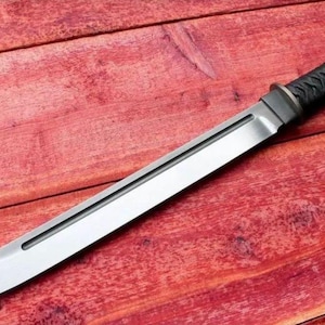 May include: A katana-style sword with a silver blade and a black, textured handle. The blade is straight with a slight curve at the tip. The handle is wrapped in a black, woven material. The sword rests on a red wooden surface.