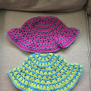 May include: Two crocheted sun hats. One hat is pink and purple, the other is blue and yellow. Both hats have a wide brim and a mesh-like pattern. The hats are handmade and appear to be made of yarn.