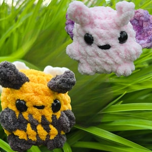 May include: Two handmade crochet plush toys. One is yellow and black, the other is pink with purple accents. Both have black eyes and smiling faces. The toys are set against a background of green grass.