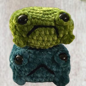 May include: Two plush frog toys, one atop the other. The top frog is a vibrant green, while the bottom is a deep teal. Both have large, black, glossy eyes and a stitched black frown, giving them a slightly sad expression. The frogs are made of a soft, textured material.