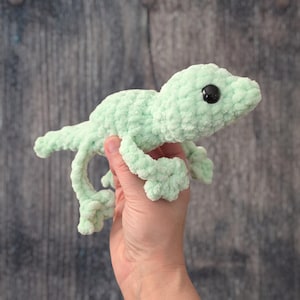 May include: A light green, plush crocheted gecko with black eyes. The handmade reptile has a textured, fuzzy appearance and is posed with its legs extended. The gecko is a soft sculpture, likely a toy or decorative item.