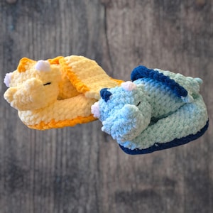 May include: Two crocheted dinosaur-shaped baby booties. One is yellow with orange trim, the other is blue with dark blue trim. Both have small white horns and closed eyes, suggesting a sleeping or relaxed state. The booties are made of soft, textured yarn.