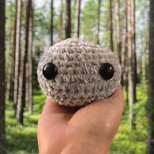 May include: A gray, crocheted plushie with two large, black button eyes. The round, fuzzy toy is held in front of a blurred forest background. The plushie is a soft, handmade item.