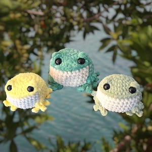 May include: Three handmade crochet frog toys in yellow, green, and light green. Each frog has black safety eyes and a white belly. The frogs are displayed against a blurred background of water and foliage.