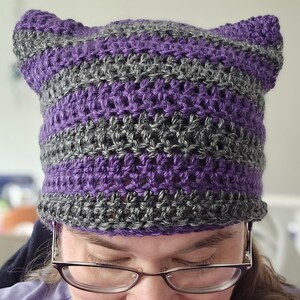 May include: A handmade crochet hat in a cat ear design. The hat features alternating horizontal stripes of purple and gray. The hat is made of a textured yarn and has a folded brim.
