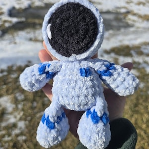 May include: A light blue crocheted astronaut doll with a black helmet and blue accents. The plush toy has a round head, arms, and legs. The doll is held in a hand against a blurred outdoor background.