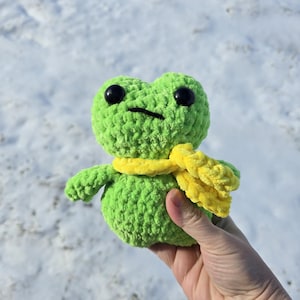 May include: A handmade, green crochet frog with black eyes and a yellow scarf. The frog is held in a person's hand against a snowy background. The frog's body is round, with small arms and legs.