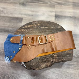 Op de afbeelding: Bruin leren riem met een denim accent. De brede riem heeft een zilveren gesp en is versierd met messing klinknagels. Het denim gedeelte is gemaakt van blauwe jeansstof.