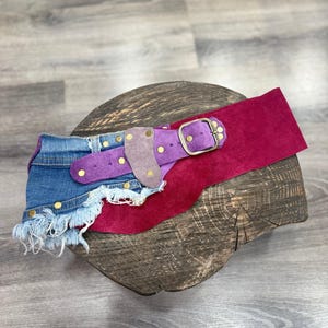 Op de afbeelding: Een veelkleurige riem met een denim gedeelte, paars suède en een magenta suède gedeelte. De riem heeft goudkleurige klinknagels en een goudkleurige gesp. Het denim gedeelte heeft een gerafelde rand.