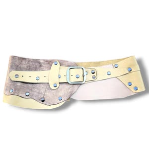 Può includere: Una cintura larga che combina pelle testurizzata color argento metallizzato e pelle liscia color crema. La cintura presenta una fibbia rettangolare in metallo argentato ed è decorata con numerosi rivetti in metallo argentato su tutta la superficie.