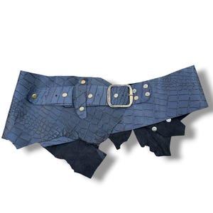 Puede incluir: Un cinturón ancho de color azul marino con un patrón texturizado de piel de cocodrilo, una hebilla rectangular de tono latón y múltiples tachuelas metálicas. El cinturón tiene un diseño asimétrico con bordes dentados y superpuestos.