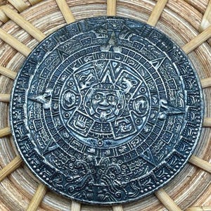 May include: A detailed, silver-colored Aztec calendar medallion. The circular design features intricate carvings of glyphs, figures, and symbols. The medallion is set against a woven, light brown background, highlighting the detailed craftsmanship of the piece.