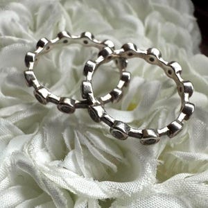 May include: Two silver rings with a repeating pattern of small, dark-colored stones. The rings are displayed on a bed of white, ruffled fabric, creating a contrast between the shiny metal and the soft background. The rings are likely jewelry.