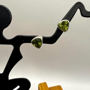 May include: A pair of stud earrings featuring triangular-cut, lime green peridot gemstones set in polished silver metal. The earrings are displayed on a black, abstract-shaped stand that holds each stone securely in place.