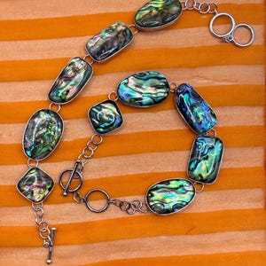 May include: Abalone shell necklace with iridescent green, blue, and purple hues. The necklace features oval and rectangular abalone shell pieces set in silver-toned metal, connected by a chain with a toggle clasp.